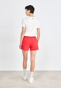 T-shirt bianca a maniche corte con logo verde, abbinata a pantaloni rossi con una tasca sul retro. Il modello indossa sneakers bianche e calzini grigi.