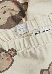 Un tissu crème présente un motif de visages de singe. Taille élastique avec une fermeture à bouton et une étiquette en textile attachée. Texture lisse.