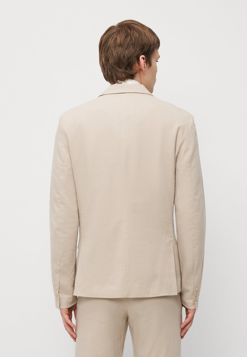 Blazer en lin beige avec une forme structurée, des revers à cran, et des manches à quatre boutons, présentant un tissu texturé et des fentes latérales.