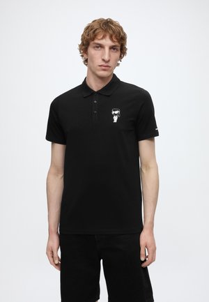 Young man with short curly hair wearing a black polo shirt with a small astronaut graphic on the left chest and black shorts.