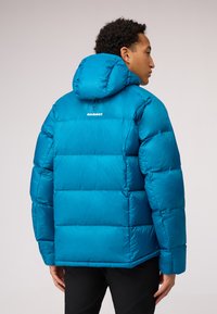 Padded blue jacket with a hood, featuring a quilted design and the "MAMMUT" logo on the back. Smooth, shiny texture with elastic cuffs.