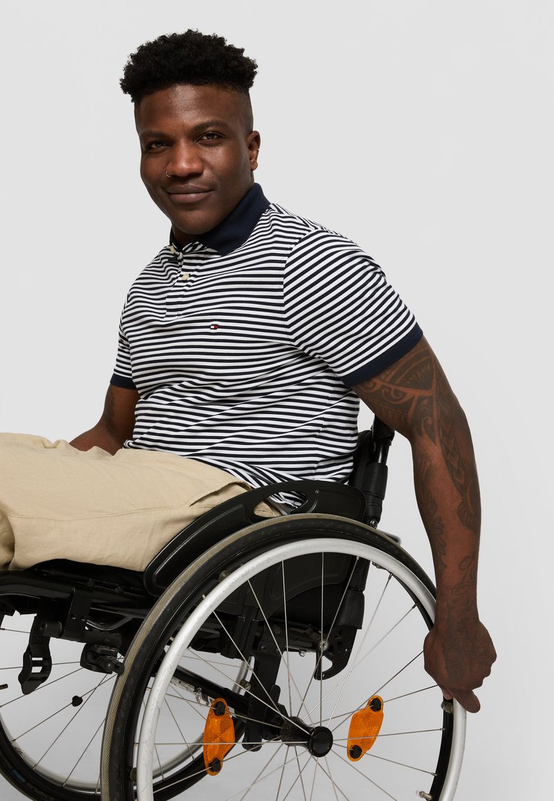 Striped navy and white polo shirt, khaki pants, seated in a black wheelchair with orange accents on the wheels. Short curly hair and tattooed arm.
