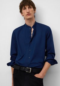 Marineblauw button-up shirt met een mandarin collar, lange mouwen en manchetten met één knoop. Gepaard met een zwarte riem en donkere broeken.