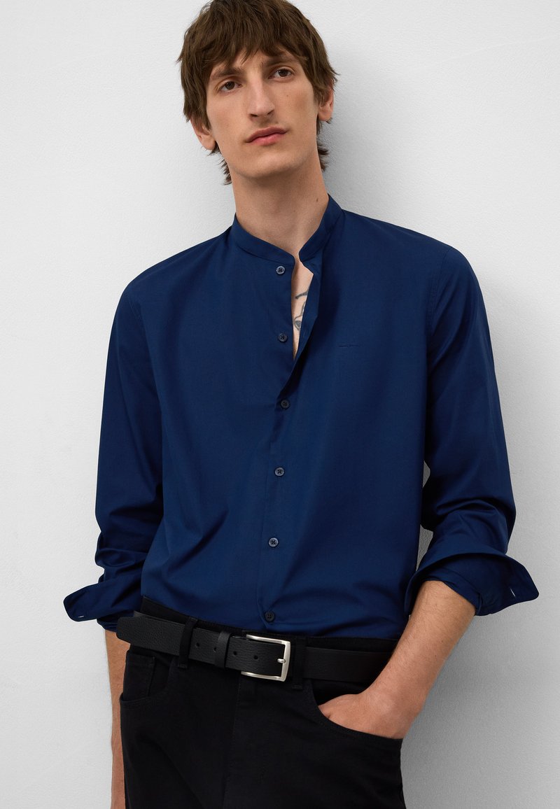 Marineblauw button-up shirt met een mandarin collar, lange mouwen en manchetten met één knoop. Gepaard met een zwarte riem en donkere broeken.
