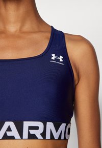 Mornarska sportska crop majica izrađena od glatkog materijala. Ima Under Armour logo u bijeloj boji i crni pojas s natpisom "ARMOUR" u bijelom slovu.