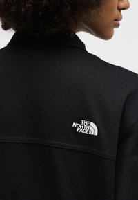Personne aux cheveux bouclés portant une veste noire avec le logo The North Face sur l'épaule arrière droite.