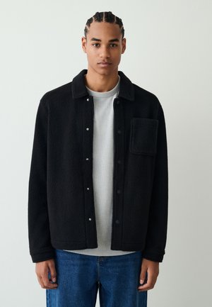 Nuori mies, jolla on letitetyt hiukset ja joka pukeutunut mustaan fleece-takkiin harmaan paidan ja sinisten farkkujen päällä, seisoo yksinkertaisen vaalean taustan edessä.