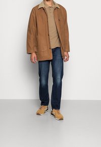 Brun bomullsjacka med en beige krage, kombinerad med en beige polo, mörkblå jeans och ljusbruna sneakers med mörka detaljer.