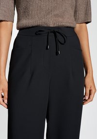 Pantaloni neri in un tessuto morbido e liscio. Presentano una vita con cordoncino, due tasche laterali e una vestibilità rilassata. Design minimalista con linee pulite.