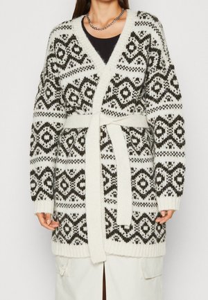 Femme portant un cardigan en maille à motif noir et blanc avec une ceinture nouée à la taille et un collier chaîne argenté.