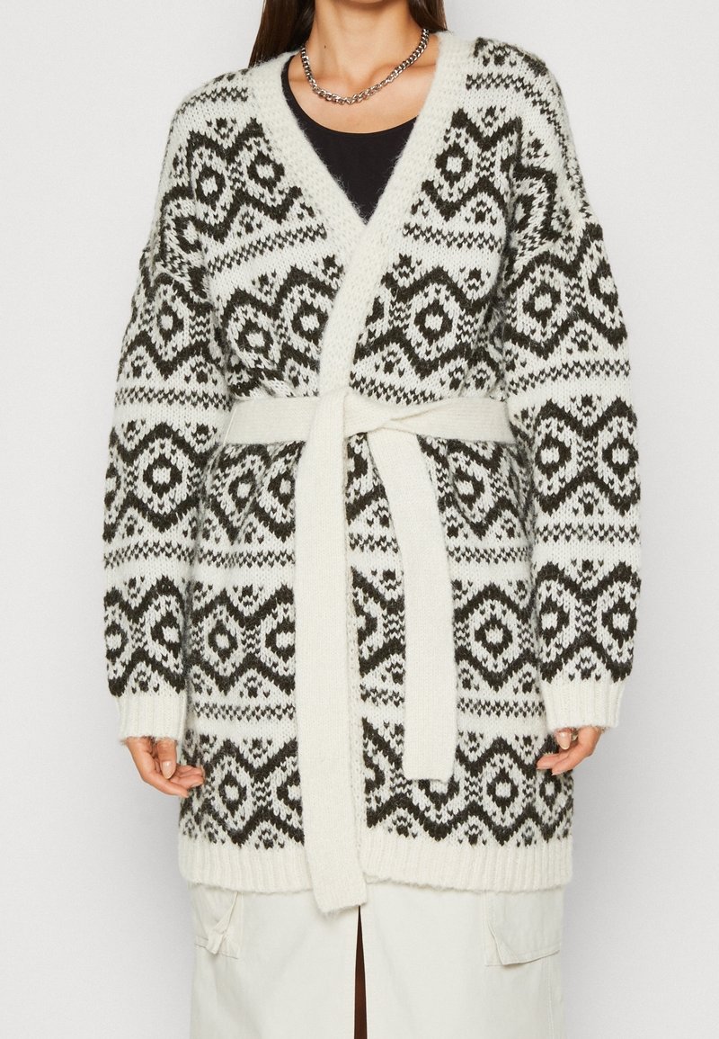 Femme portant un cardigan en maille à motif noir et blanc avec une ceinture nouée à la taille et un collier chaîne argenté.