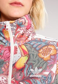 Kurtka zip-up w wielokolorowy wzór kwiatowy z wysokim kołnierzem, z motywem tukana i logo Adidas na dolnym lewym boku.