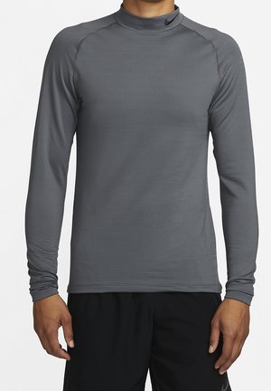 Mężczyzna w dopasowanej, długorękawowej, szarej sportowej koszulce z wysokim kołnierzem i małym czarnym logo przy szyi, w zestawie z czarnymi szortami.