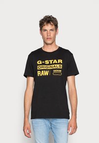 Camiseta negra con texto amarillo que presenta "G-STAR RAW" y "DENIM", de manga corta, cuello redondo, tela suave, combinada con jeans azul claro.