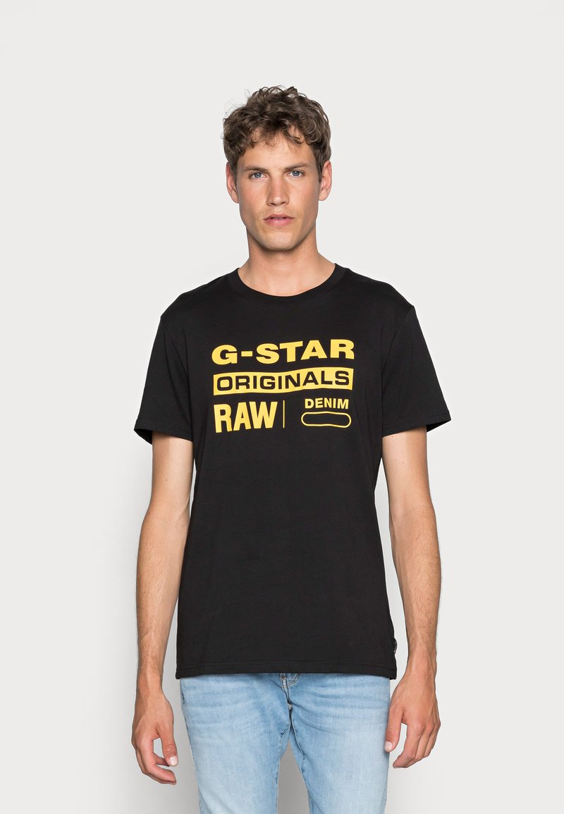 Camiseta negra con texto amarillo que presenta "G-STAR RAW" y "DENIM", de manga corta, cuello redondo, tela suave, combinada con jeans azul claro.