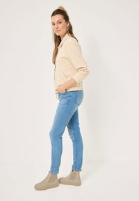 Veste zippée beige avec des poignets et un ourlet côtelés, associée à un jean skinny bleu clair. Bottines beiges avec panneaux élastiques sur les côtés.