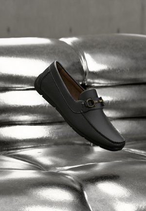 Mocassin en cuir noir avec détail de boucle métallique flottant au-dessus d'un canapé en cuir argenté brillant.