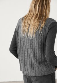 Grauer Strickpullover mit einem strukturierten Zopfmuster auf dem Rücken und gerippten, langen Ärmeln, kombiniert mit dunklen Jeans. Natürliches Haar fällt über die Schulter.