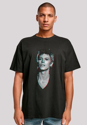 Man with short hair wearing black oversized T-shirt featuring a distorted vintage portrait of a person with a cross necklace.