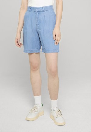 Persona vistiendo pantalones cortos plisados de talle alto color azul claro, calcetines blancos y zapatillas blancas, de pie frente a un fondo liso gris claro.