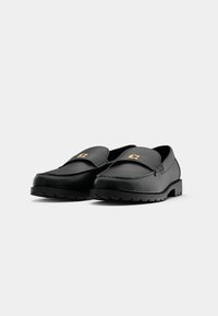 Mocassins en cuir noir avec semelles épaisses et emblème doré sur la bride, présentés côte à côte sur fond blanc.