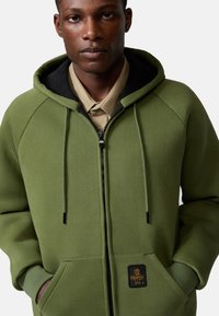 Felpa verde con zip frontale, cappuccio e maniche raglan. Presenta una fodera interna nera e un'etichetta sulla tasca frontale.