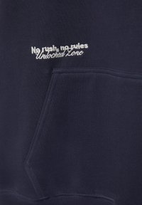 Tengerészkék szövet elejtett zsebbel és fehér hímzett szöveggel: "Nincs sietség, nincs szabály Feloldott zóna."