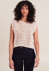 Gehaakte top met gegolfde mouwen, gemaakt van beige garen en voorzien van openwerk patronen. Gecombineerd met donkere jeans, pols versierd met een gouden armband.