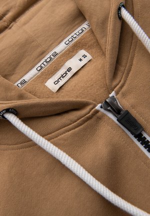 Sudadera con capucha beige con cordones blancos, cremallera negra con la etiqueta "OMBRE" y etiquetas interiores que indican la marca "ombre" y talla mediana 48.