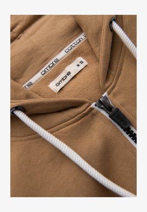 Beige hoodie with white drawstrings, black zipper labeled "OMBRE," and interior labels indicating brand "ombre" and size medium 48.