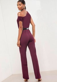 Frau in einem pflaumenfarbenen Ein-Schulter-Jumpsuit mit einem schwarzen Gürtel, goldenen Ohrringen und nudefarbenen High Heels, stehend vor einer weißen Wand.