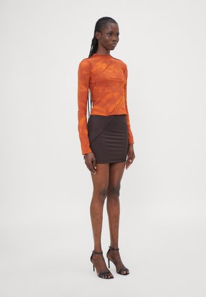 Orangenes Langarmoberteil mit strukturiertem Muster, kombiniert mit einem taillierten braunen Minirock mit diagonalen Nahtdetails und schwarzen Riemchenheels.