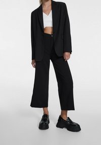 Zwarte op maat gemaakte blazer over een witte crop top, gecombineerd met brede zwarte broek en stevige zwarte loafers. Vlotte texturen en gestructureerde vormen.