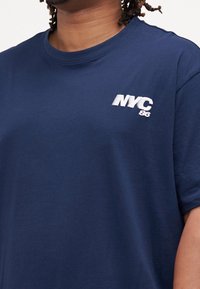 Tmavě modré bavlněné tričko s kulatým výstřihem a krátkými rukávy, s bílým vyšívaným logem "NYC 86" na levé straně hrudi. Měkká textura.