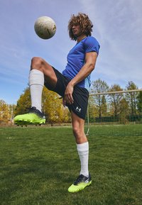 Młoda osoba w niebieskiej koszulce sportowej i czarnych szortach balansuje piłką nożną na kolanie, mając na sobie jaskrawozielone korki i białe skarpety.