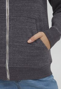 Mano en el bolsillo de una sudadera con cremallera de color gris oscuro, combinada con jeans azul claro, mostrando la cremallera y la textura de la tela.