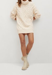 Kerman väri villapaita mekkona, jossa on korkea kaulus ja lyhyet hihat, yhdistettynä beigeihin nilkkabootseihin. Tekstuuriset kuviot kauttaaltaan.