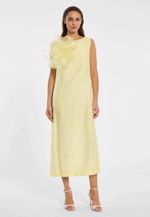 Vrouw draagt een mouwloze, lichtgele midi-jurk met een groot doorschijnend bloemdetail op de linkerschouder en witte sandalen met hoge hakken.