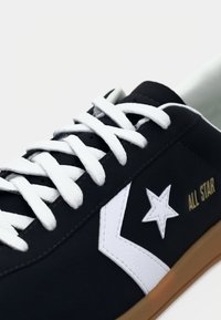 Black sneaker with white laces, white chevron and star logo, gold "ALL STAR" text, and light brown sole on a white background.