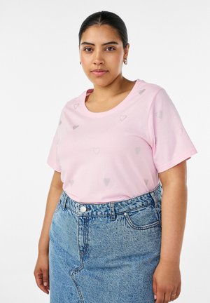 Kobieta stoi przodem, ubrana w jasnoróżowy t-shirt z małymi wzorami serduszek i niebieską, wysoką w talii dżinsową spódnicę, na białym tle.