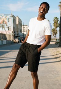 Mosolygó férfi fehér pólóban és fekete rövidnadrágban áll a napsütötte városi utcán, háttérben pálmafák és egy falfestmény látható.