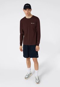 Bruine lange mouwen shirt met klein logo, marineblauwe korte broek, grijze en witte sneakers en een marineblauwe pet, staande op een effen achtergrond.
