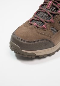 Primo piano di una scarpa da trekking marrone con suola robusta, lacci a strisce rosa e dettagli in tessuto a rete su sfondo bianco.