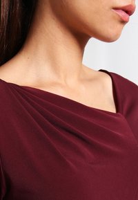 Burgundy dress featuring a draped neckline and smooth texture, showcasing subtle pleats along the shoulders.