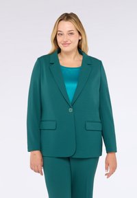 Žena s blond vlasy v tyrkysovém saku přes odpovídající tyrkysový saténový top a tyrkysové kalhoty, stojící proti bílému pozadí.