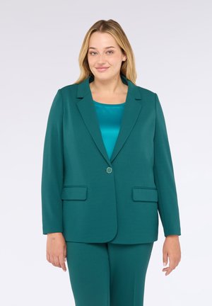 Donna con capelli biondi che indossa un blazer teal sopra una camicia di satin teal abbinata e pantaloni teal, in piedi contro uno sfondo bianco.