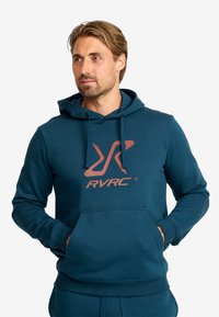 Turchinsydd hoodie iverkledt i mjukt materiale med kengurulomme og stor RVRC-logo i rustfarge foran. Klassisk passform med snor i hetten.