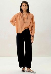 Blouse orange clair avec encolure froncée, associée à un pantalon large taille haute noir et des ballerines argentées métalliques. Collier en perles en détail.
