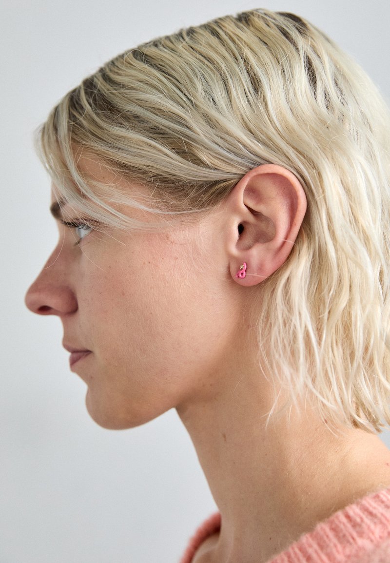 Cercei stud în formă de flamingo roz cu o finisare lucioasă, purtați pe o coafură scurtă de blonda deschisă. Urechea are o textură simplă și naturală.
