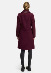 Manteau en mélange de laine bordeaux avec une coupe ajustée, grand col et deux détails accentués à l'arrière. Associé à des leggings noirs et des bottes.